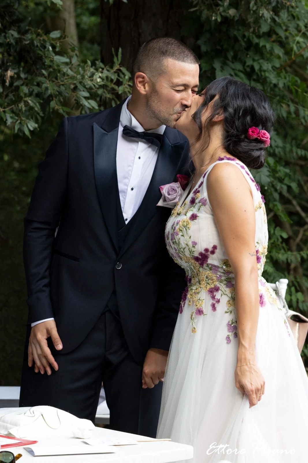 Gli sposi si baciano davanti al verde del giardino; la sposa indossa un abito con decorazioni floreali.