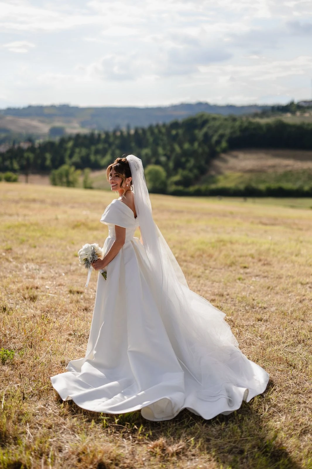 Sposa con velo e bouquet in un prato, con le colline sullo sfondo.