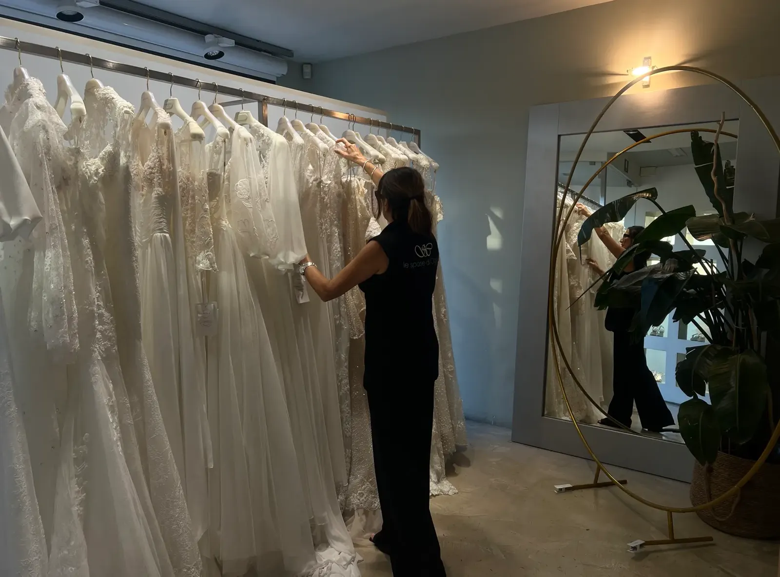 Una persona dello staff sceglie tra gli abiti da sposa appesi, riflessa nel grande specchio dell’atelier.