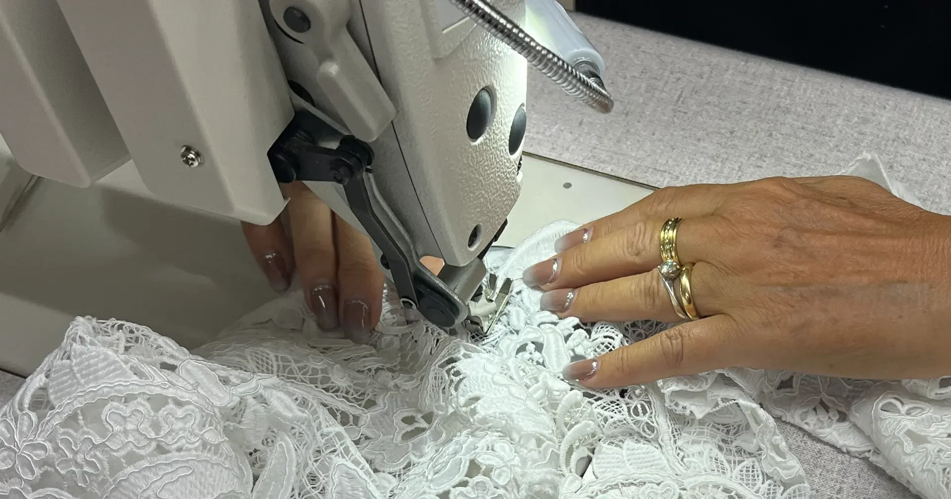 Dettaglio delle mani al lavoro su un abito in pizzo bianco alla macchina da cucire nell’atelier.