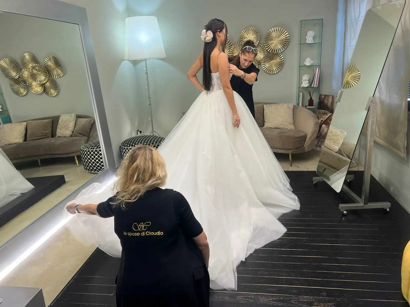 Sposa in abito con gonna ampia in tulle durante la prova, assistita dallo staff che sistema corpetto e strascico.